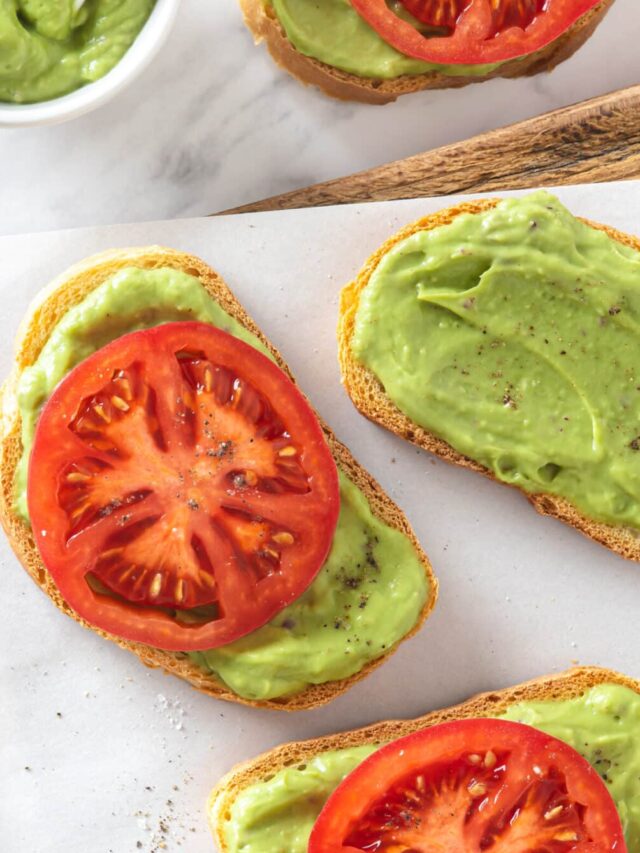 avocado toast pieces with a large tomato slice on each of them with some on a wooden board with parchment paper and a bowl of smooth avocado topping with a spoon in it.