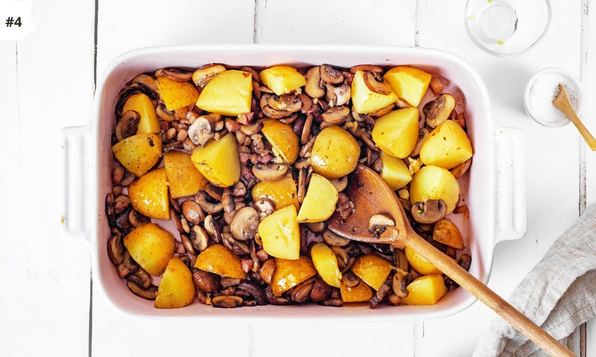 Potatoes and mushrooms ready to bake in a white baking dish with a wooden spoon and a small bowl of salt on the side.