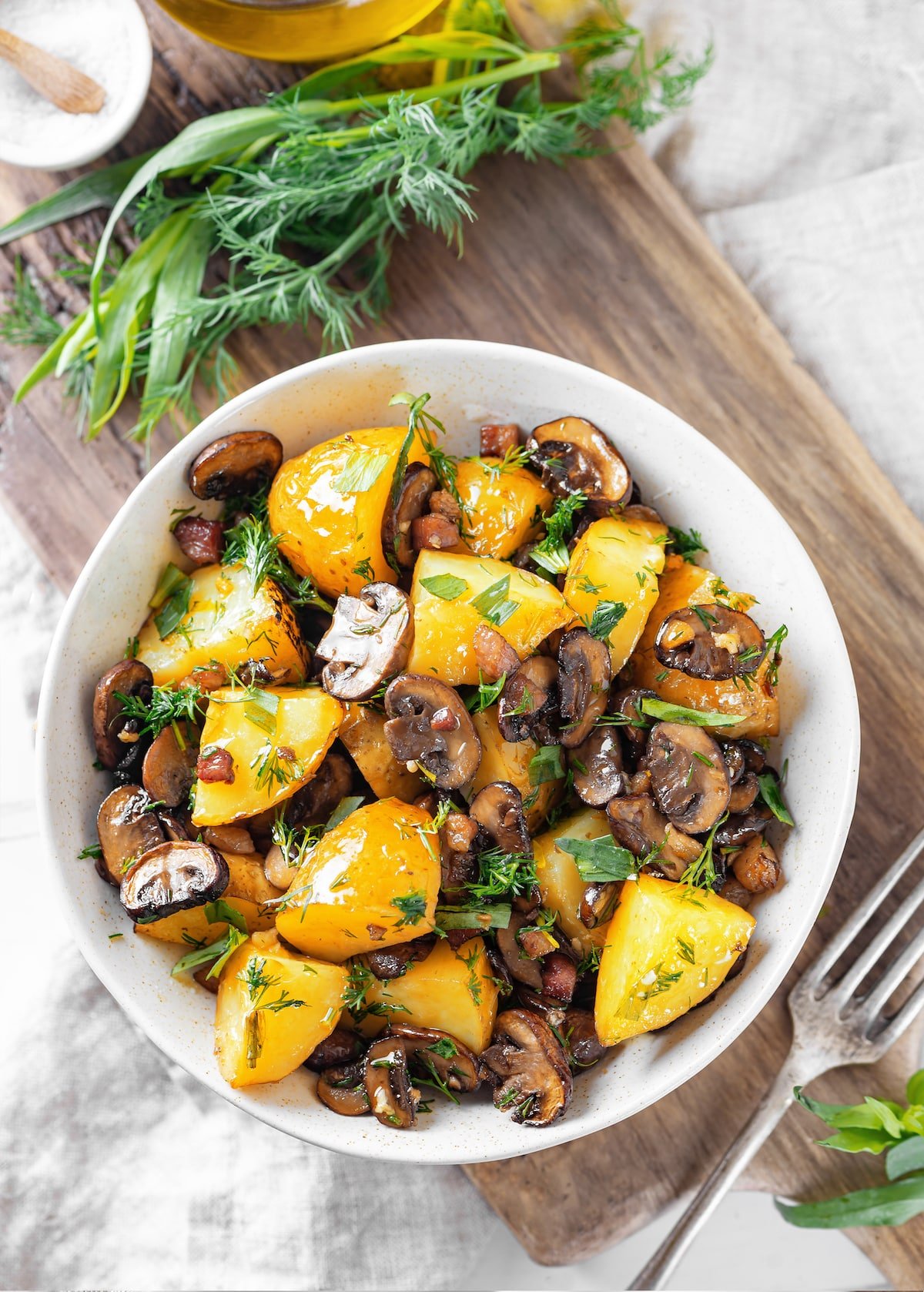 A white bowl filled with roasted potatoes, mushrooms, and pancetta, as well as greens on a wooden board with a fork on the side.