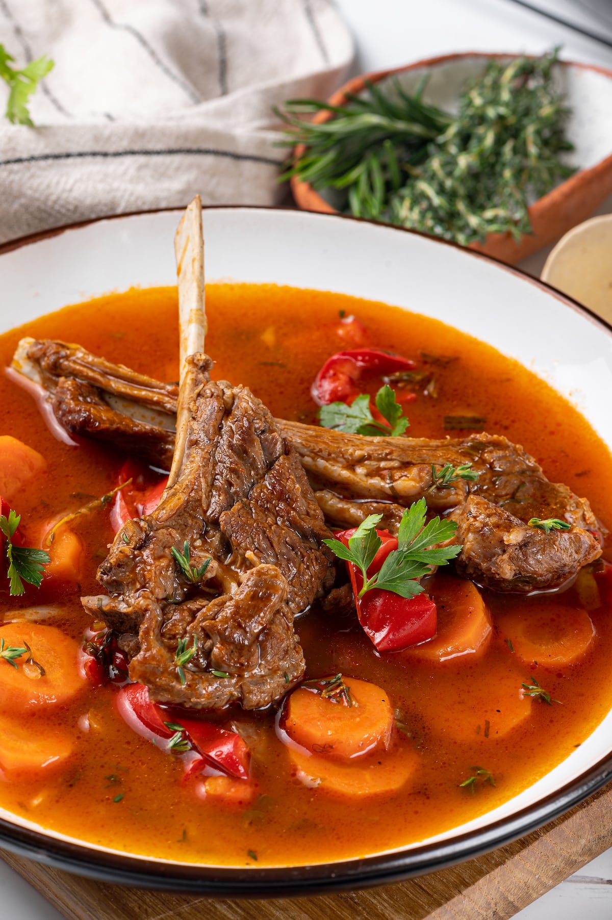 lamb chops in tomato water with herbs and chopped vegetables in a white bowl.