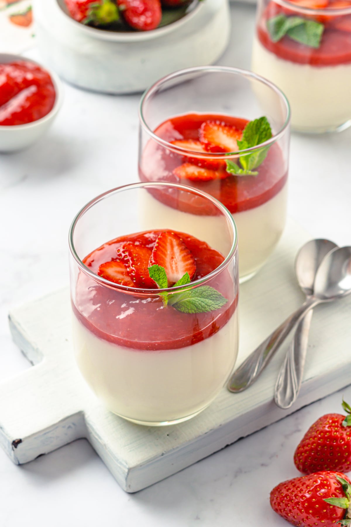 a couple glasses of strawberry panna cotta with strawberry slices and mint on top, with two spoons set to the side on a serving board