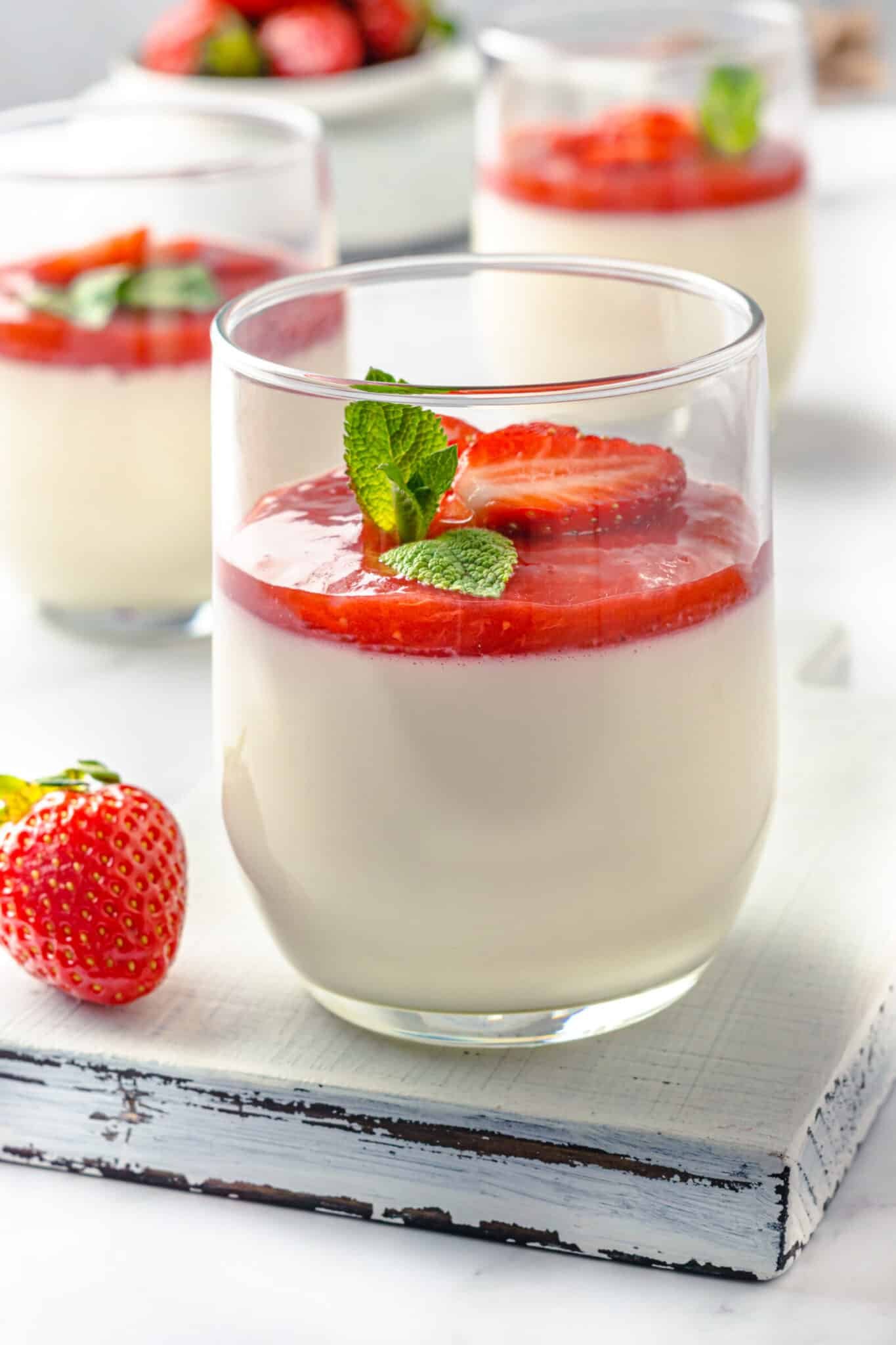 panna cotta in a glass with strawberry jam on top and sprigs of mint leaves and slices fresh strawberries for garnish.