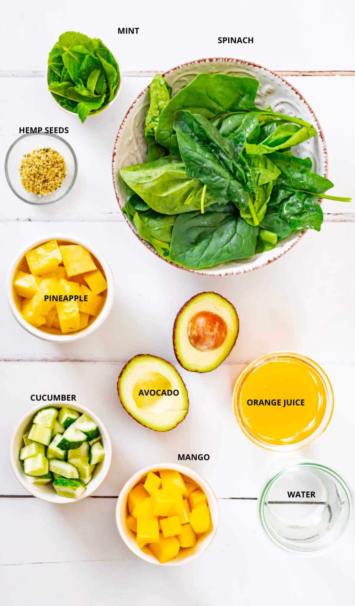 citrus green smoothie ingredients with labels mint spinach hemp seeds pineapple avocado cucumber mango orange juice and water.