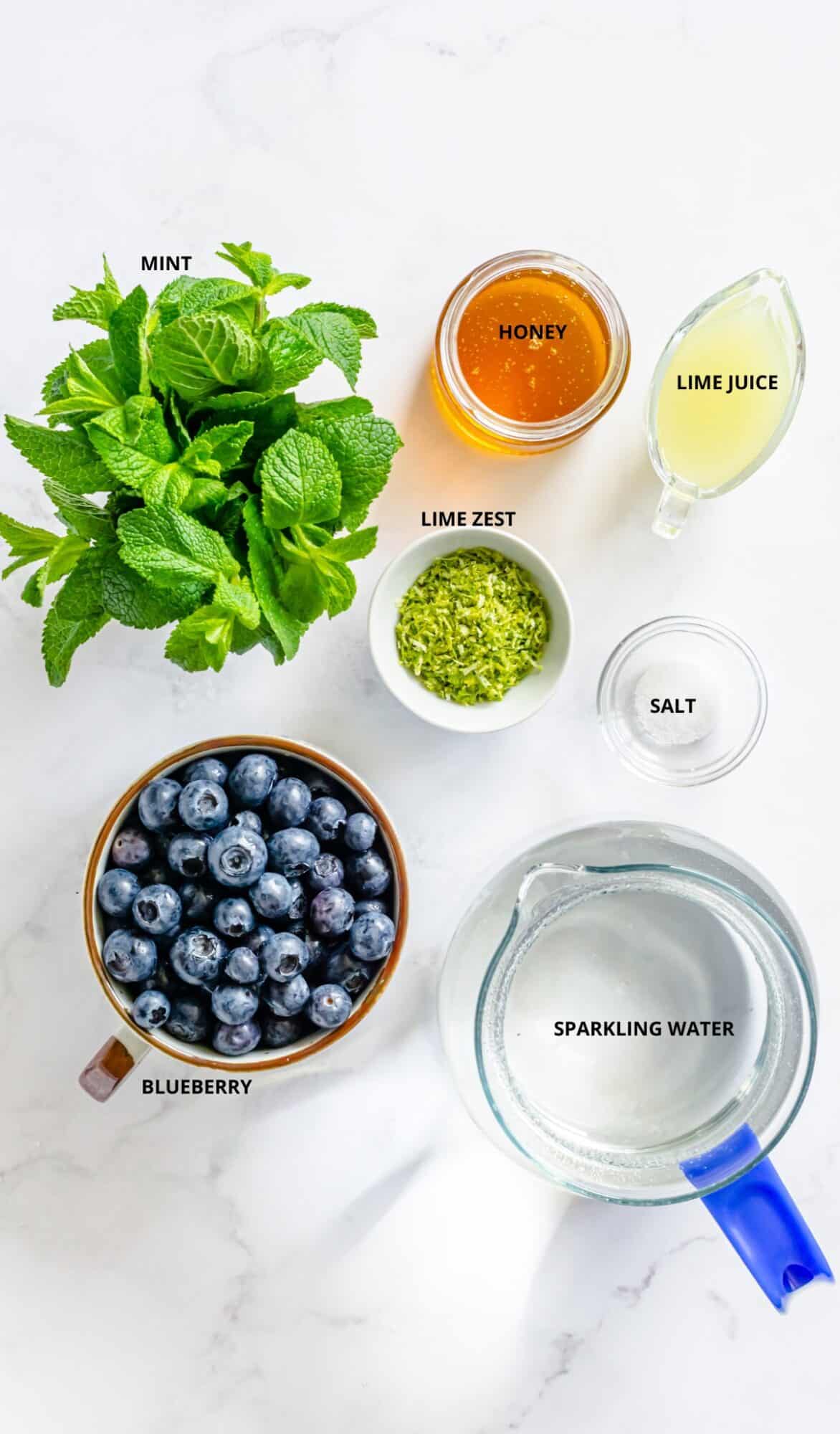 Fresh mint, honey in a jar, fresh lime juice in a clear cup, lime zest in white shallow bowl, pinch of salt in clear small bowl, sparkling water in a clear pitcher, fresh blueberries in a white and brown mug.
