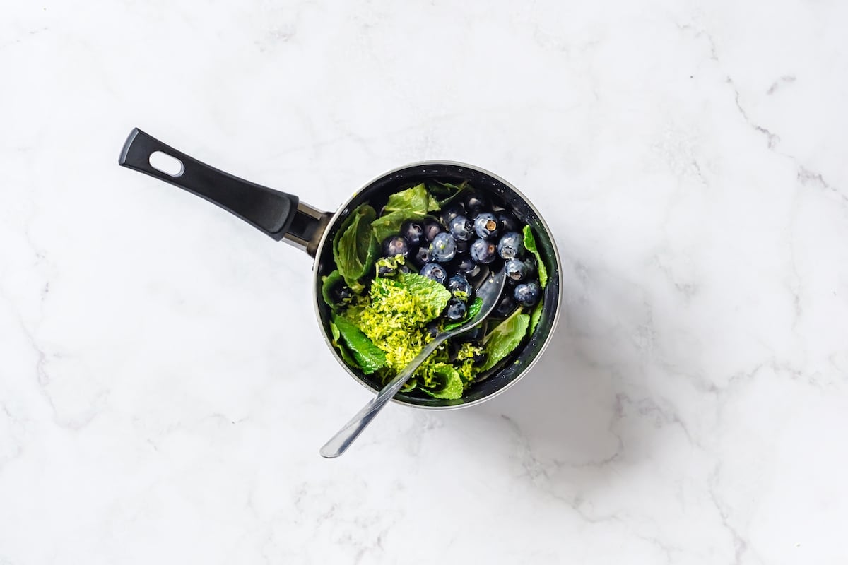 Fresh blueberries, mint, lime zest, in a black medium pan with silver tablespoon spoon.