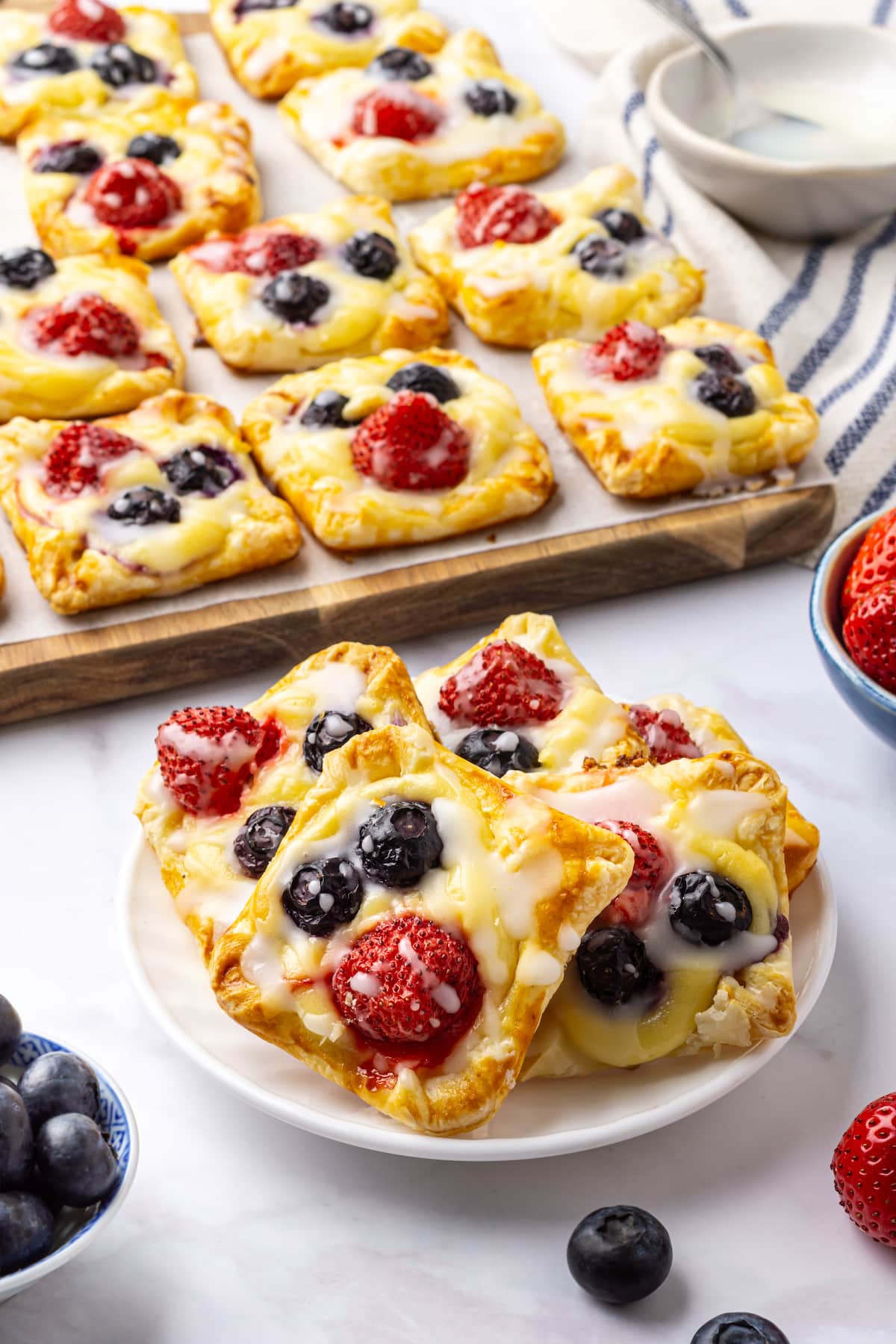 glazed cream cheese danishes with strawberries and blueberries on top lined up on a wood block in the background, as well as stacked on a white plate in the foreground.