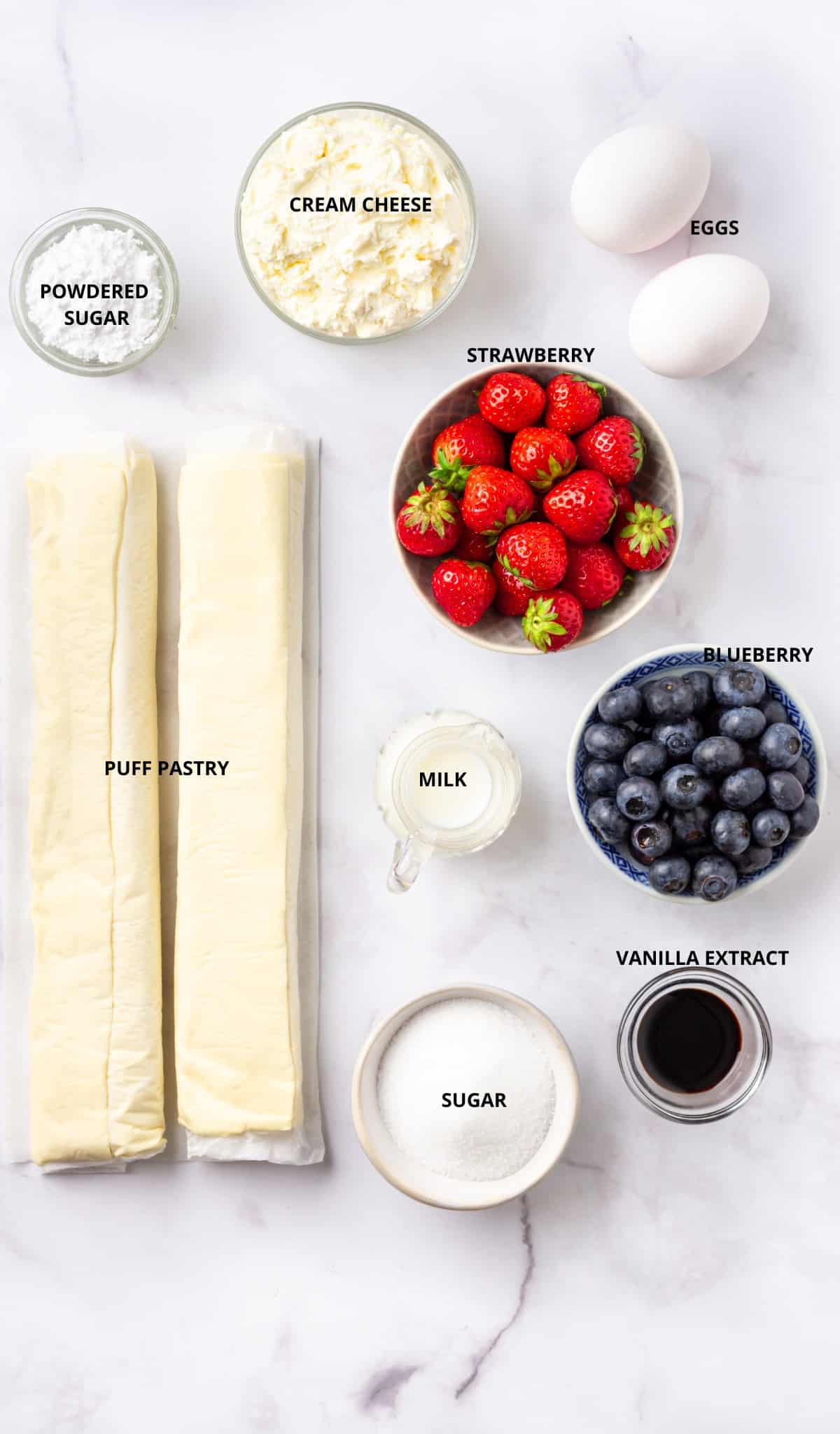 all of the ingredients for the cream cheese danish recipe on a white marble surface: cream cheese, powdered sugar, strawberries, eggs, puff pastry, milk, blueberries, vanilla extract, and sugar