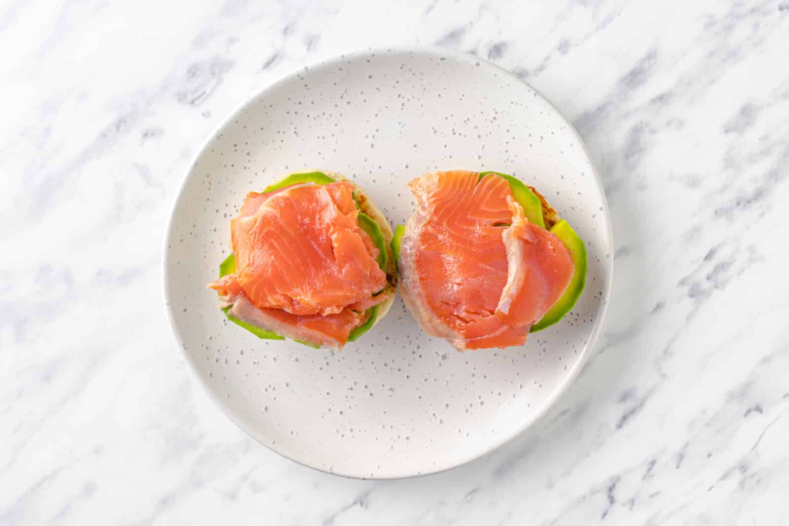 smoked salmon on top of avocado on top of two halves of english muffins, all on a plate