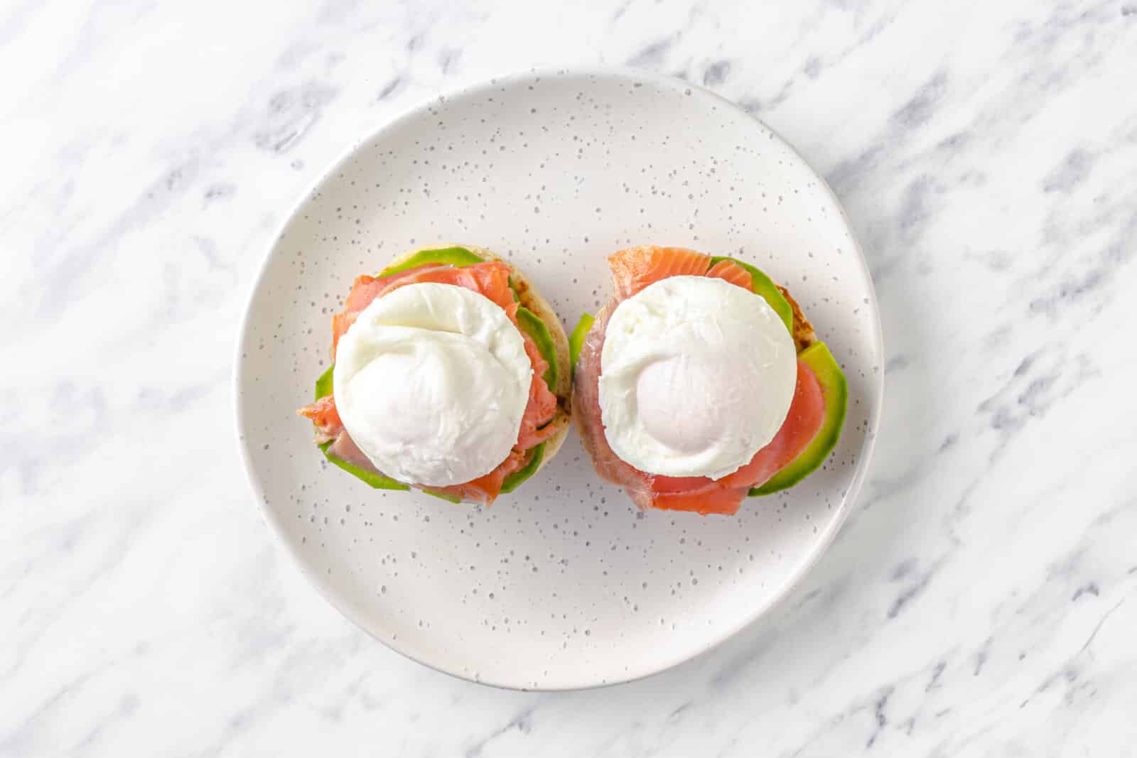 two halves of english muffins on a plate with avocado, smoked salmon, and poached eggs on top