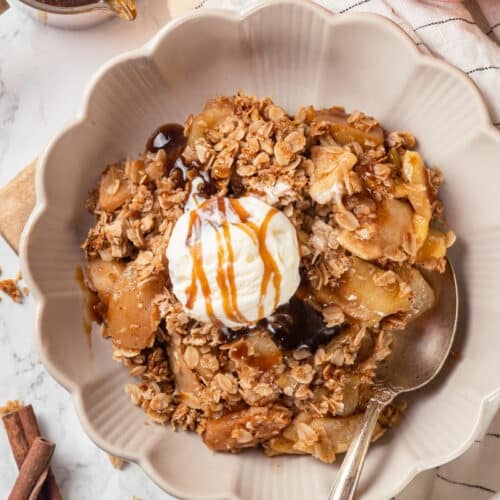 A bowl of apple crisp served with a scoop of ice cream and a spoon, with more apples on the side.