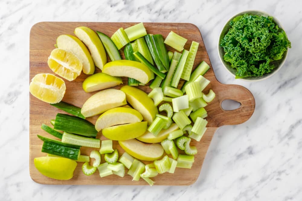 Chopped apples, cucumber, lemon, and celery on a wooden board with kale on the side in a bowl.