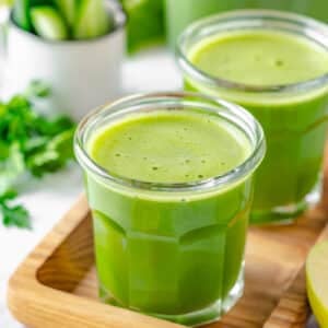 Green juice in two glasses atop a wooden tray with lemon on the side.