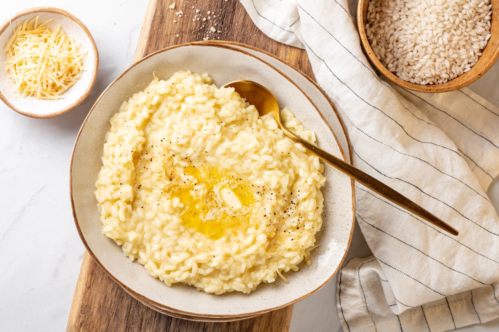 Risotto in a white bowl with melted butter and pepper on top, with a spoon on the side andmore cheese for topping.