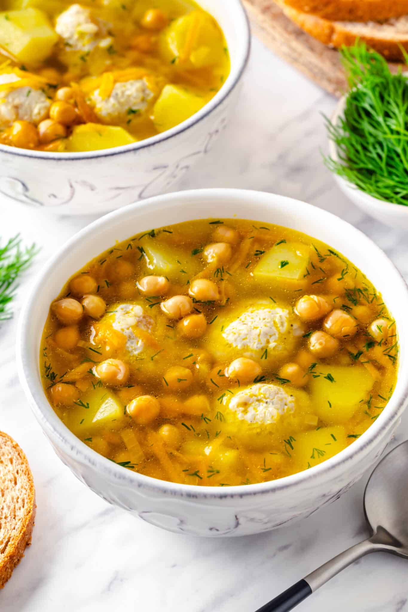 Garbanzo bean soup with meatballs and potatoes in a white bowl with a spoon on the side.