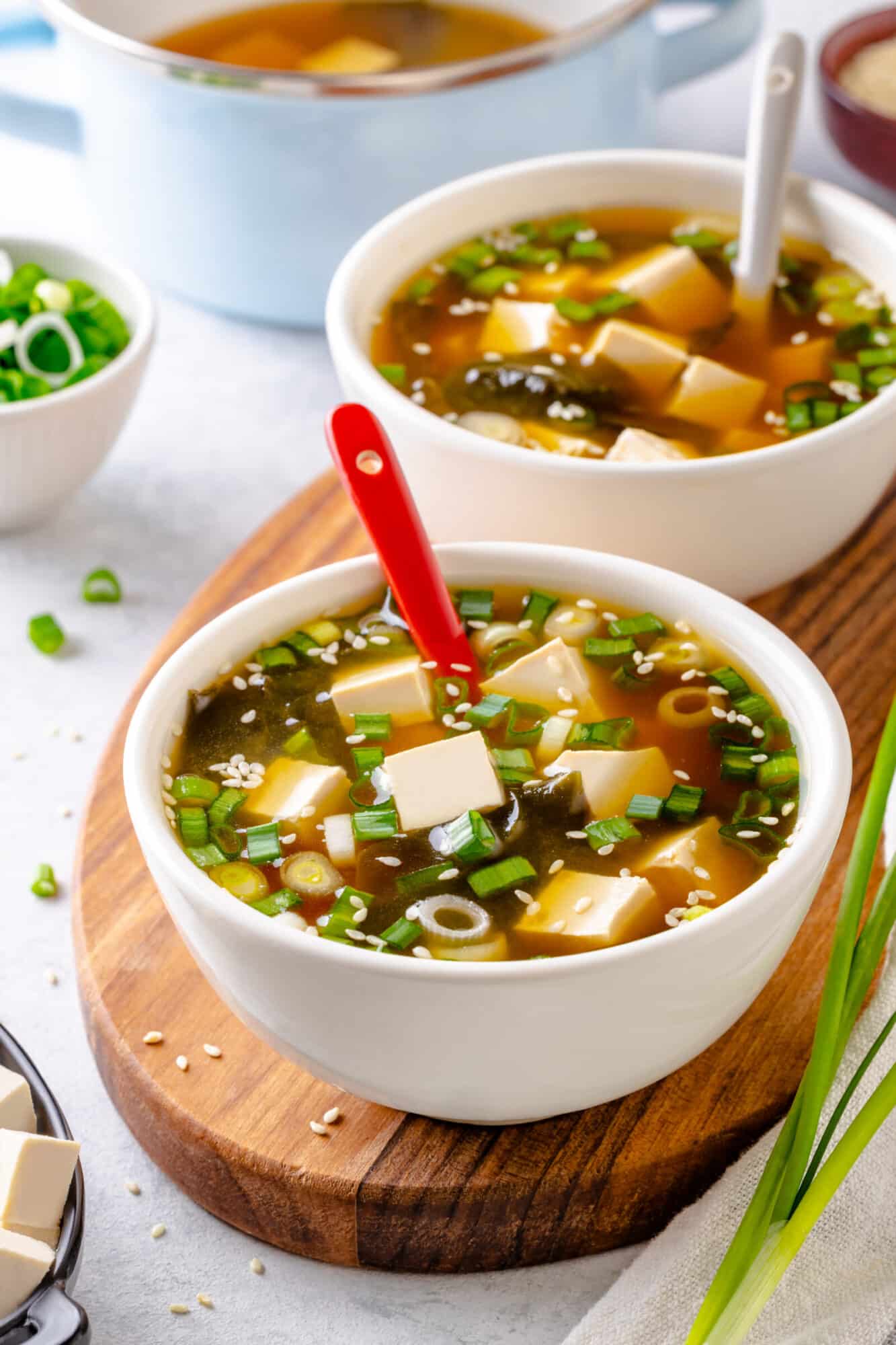 How To Make Miso Soup At Home A bowl of miso soup atop a wooden display board with red and white spoons.