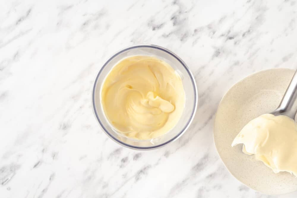 Blended egg with oil, mustard, salt, and lemon juice in a cup with the immersion blender on the side.