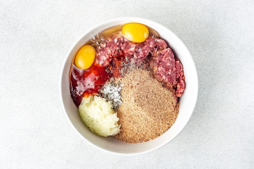 Breadcrumbs, meat, spices, egg, and onion mixed in a white bowl.