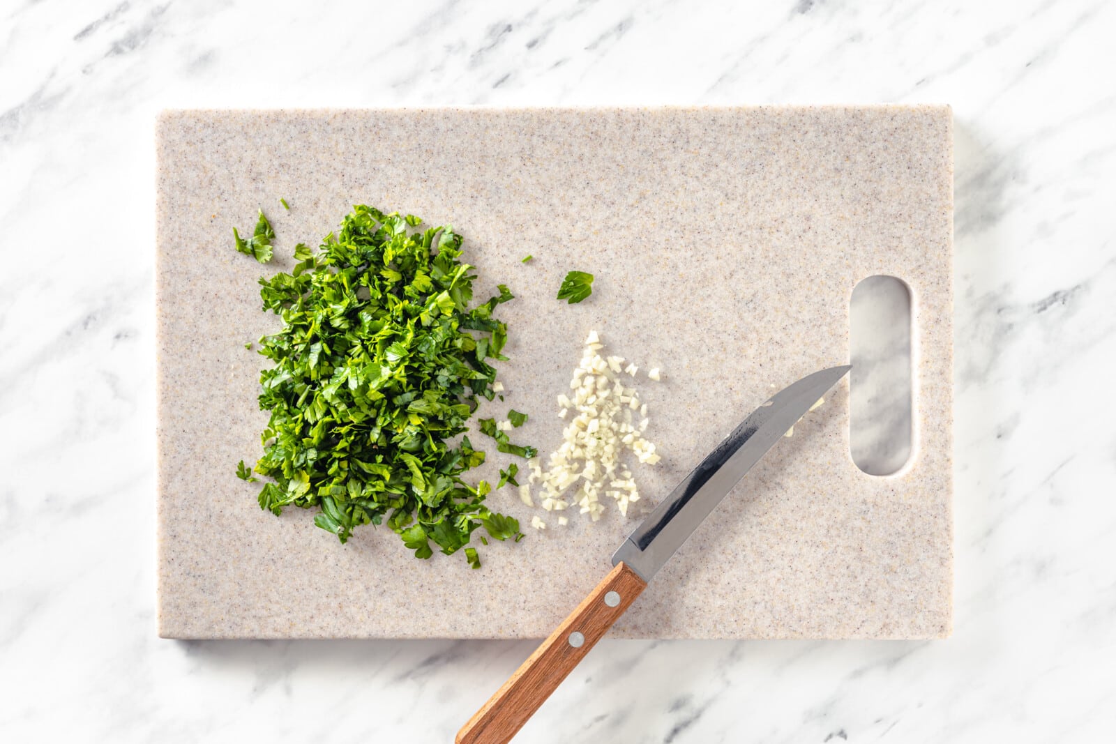Chopped parsley and minced garlic atop a cutting board with a knife.