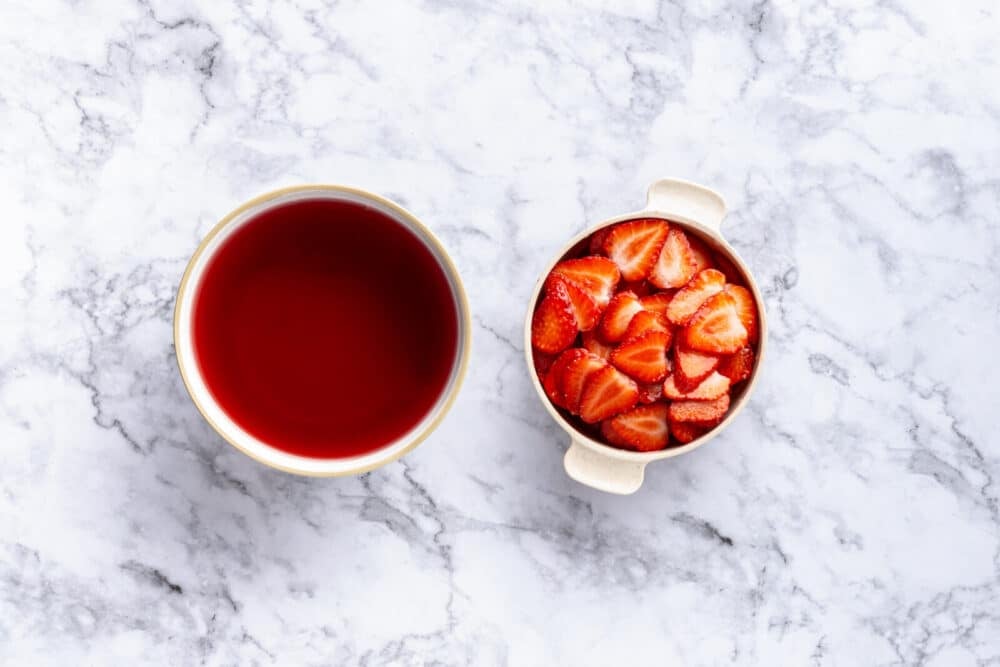Jello in a white bowl with sliced strawberries in a white bowl with handles.