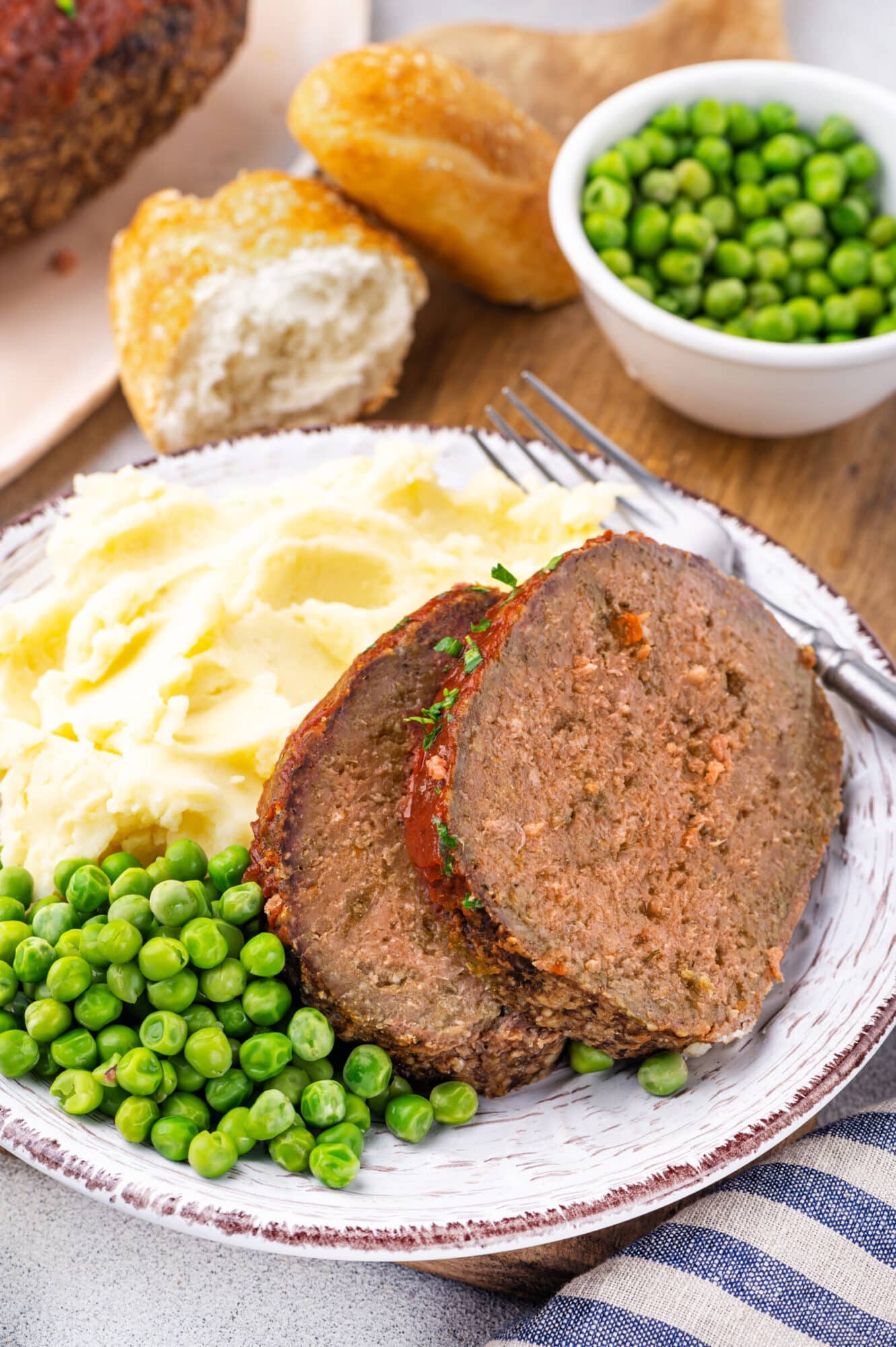Meatloaf served with peas and mashed potatoes atop a plate with bread on the side.