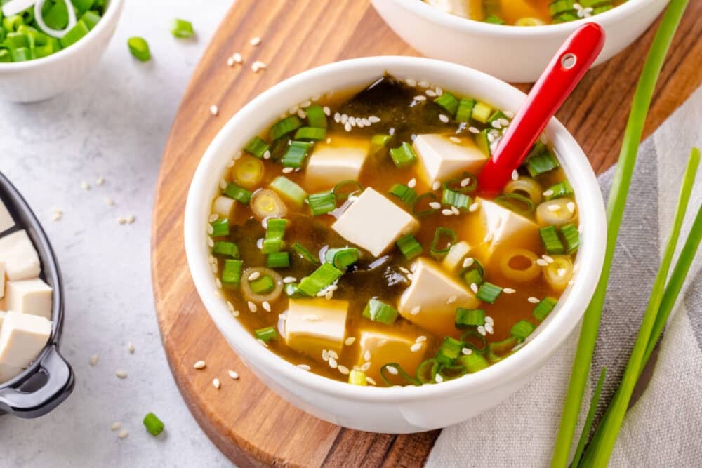 Miso soup in a large white bowl, serving atop a wooden board with green onion on the side.