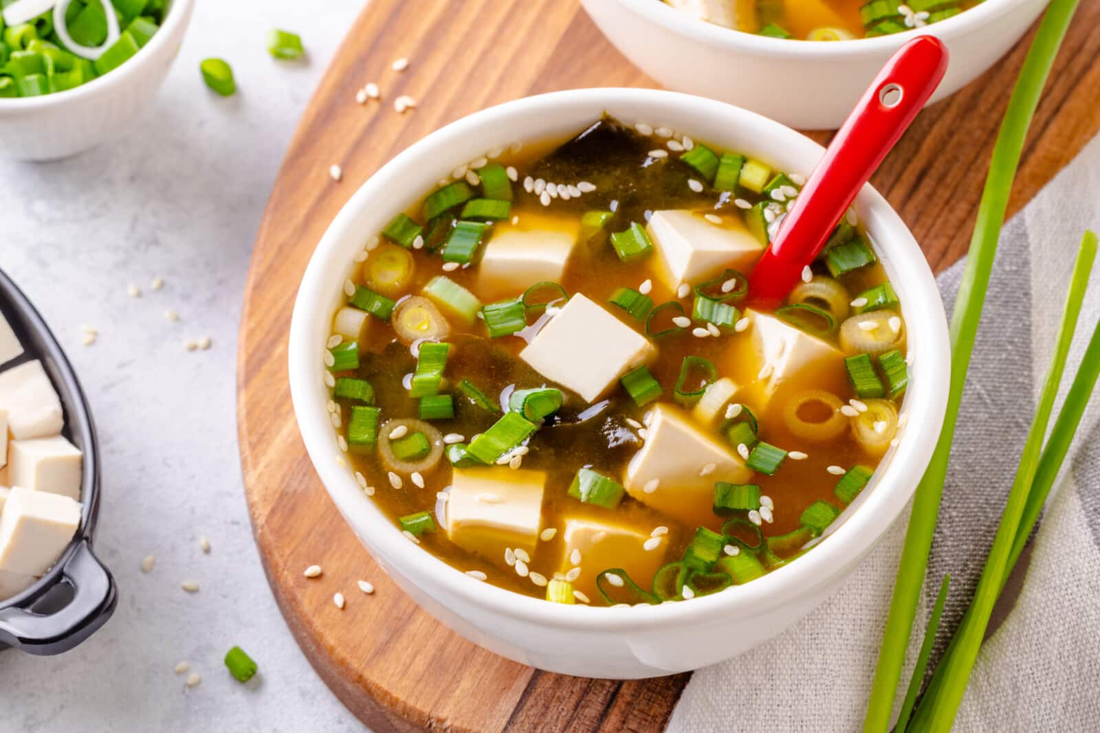 Miso soup in a large white bowl, serving atop a wooden board with green onion on the side.