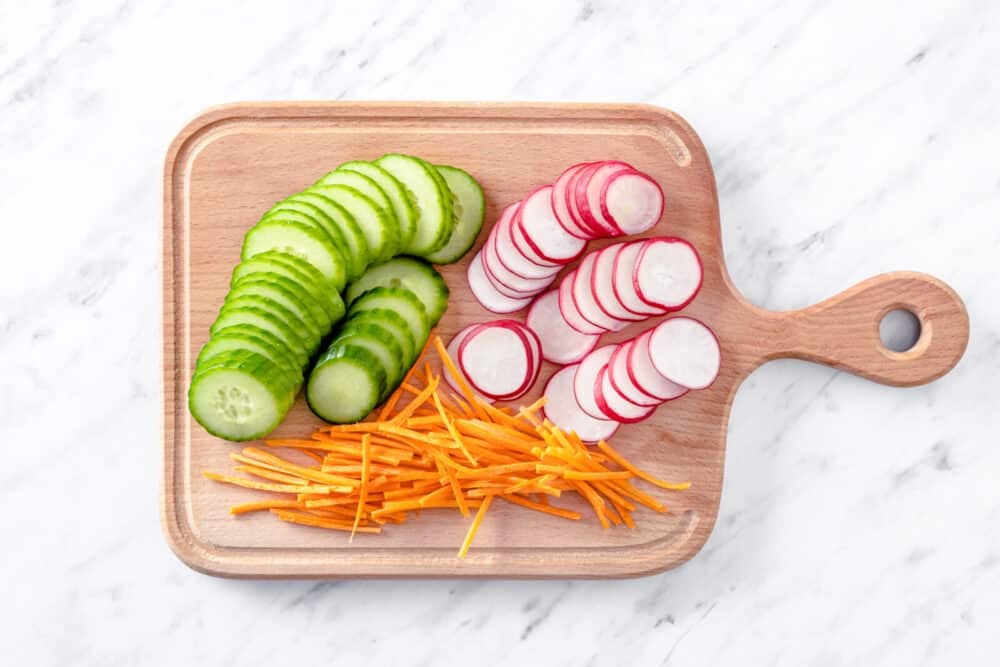 Sliced cucumber, radishes, and julienned carrots atop a wooden board.
