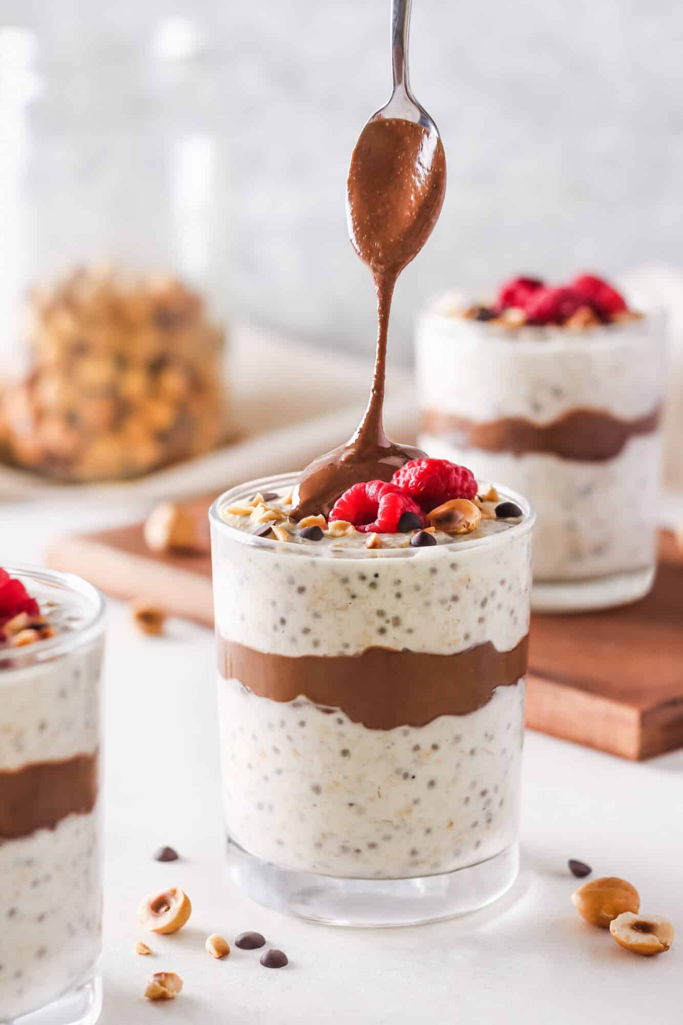 Spooning hazelnut butter atop overnight oats in a glass with berries and nuts for topping.